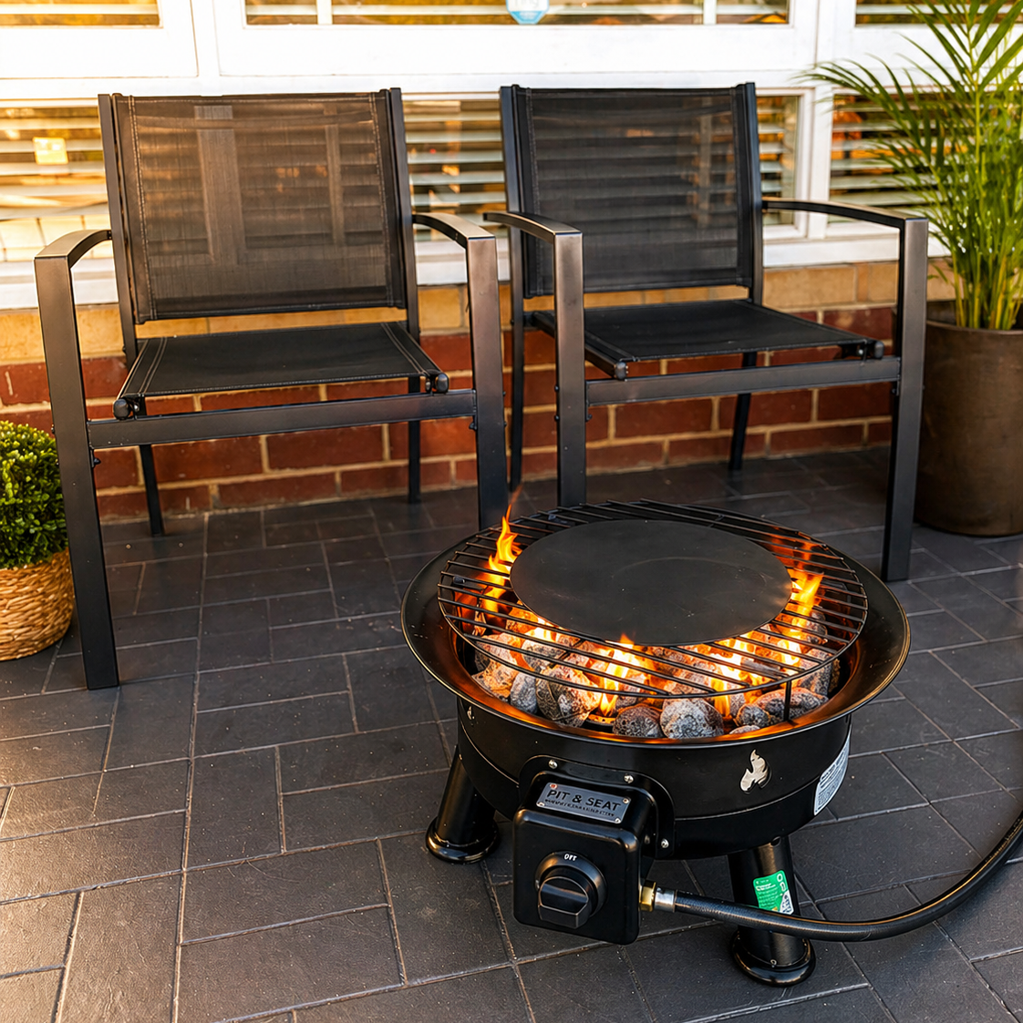 Two black chairs around a BBQ Flame on a patio with a brick wall and plants in the background.